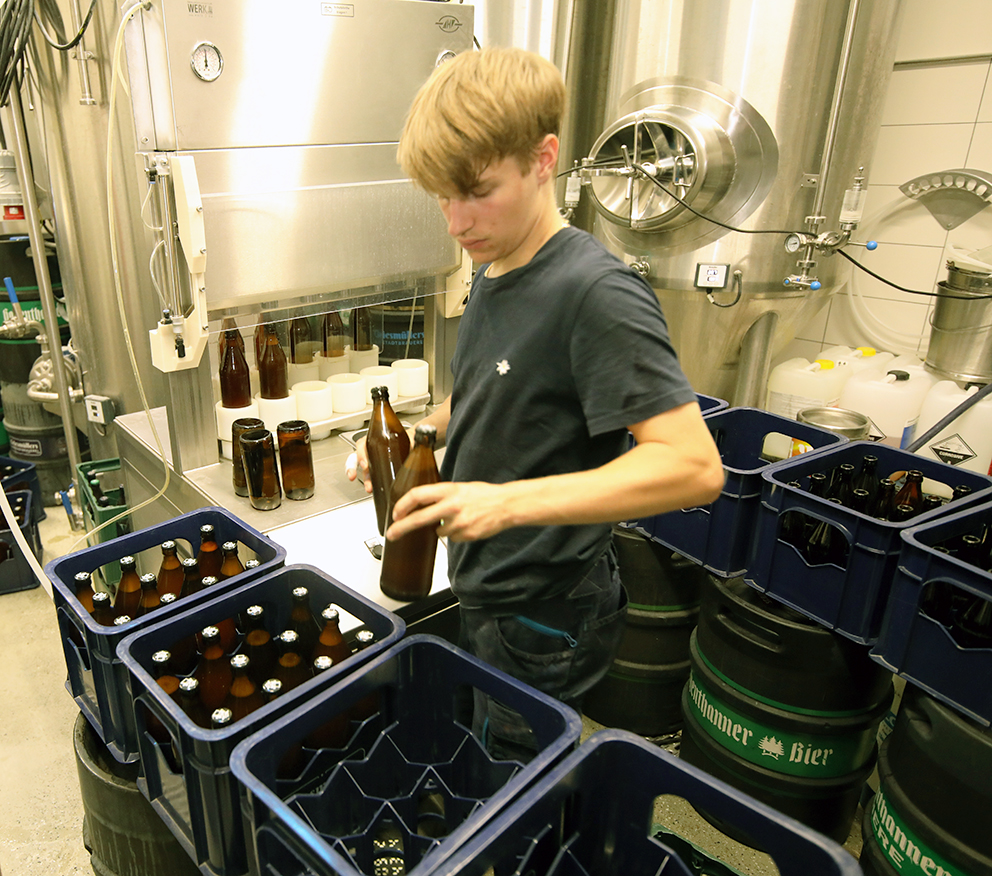 A brewer placing filled brown bottles into blue crates beside the fermentation tanks