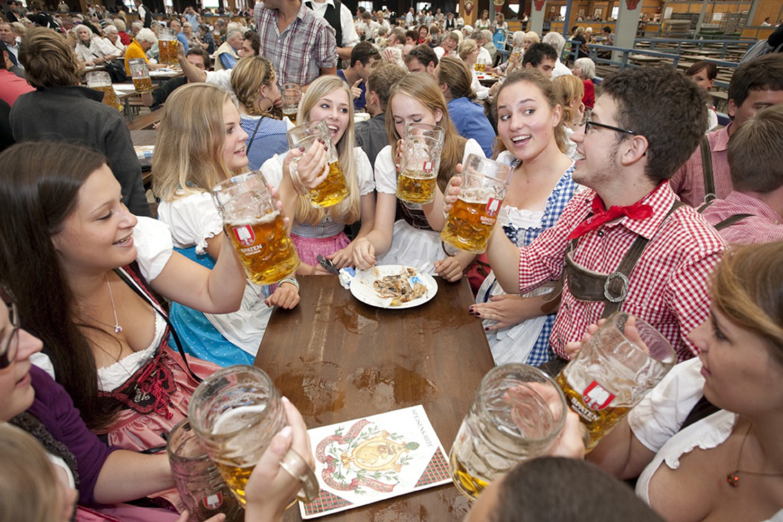 A group of friends in dirndls and lederhosen raising beer steins together at a long Oktoberfest table
