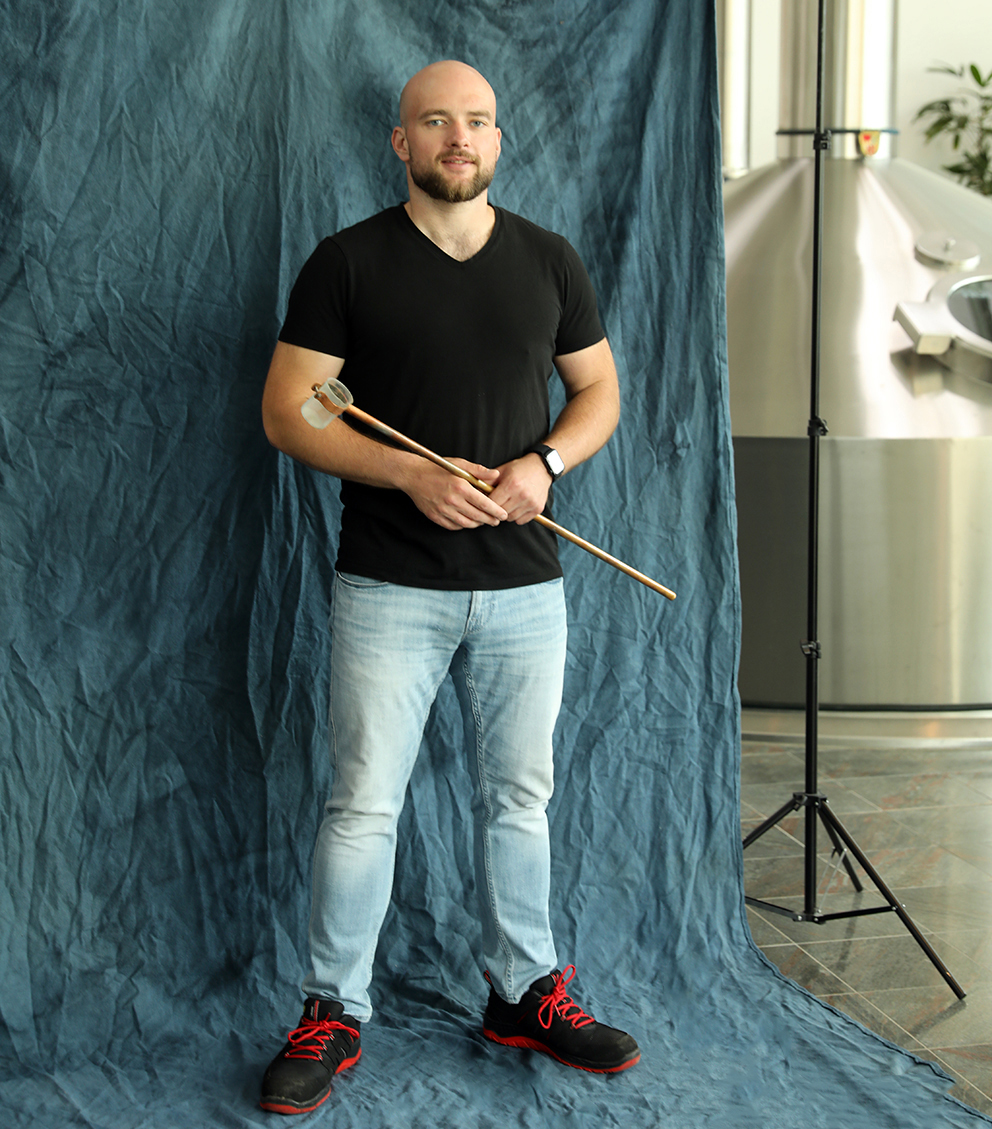 A brewer in a black shirt holding a long brewing instrument against a blue backdrop