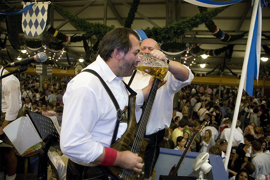 A musician in traditional dress drinking from a beer stein while performing inside an Oktoberfest tent
