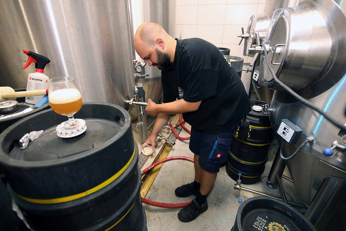 A brewer filling a keg from the fermentation tank, a full glass standing nearby