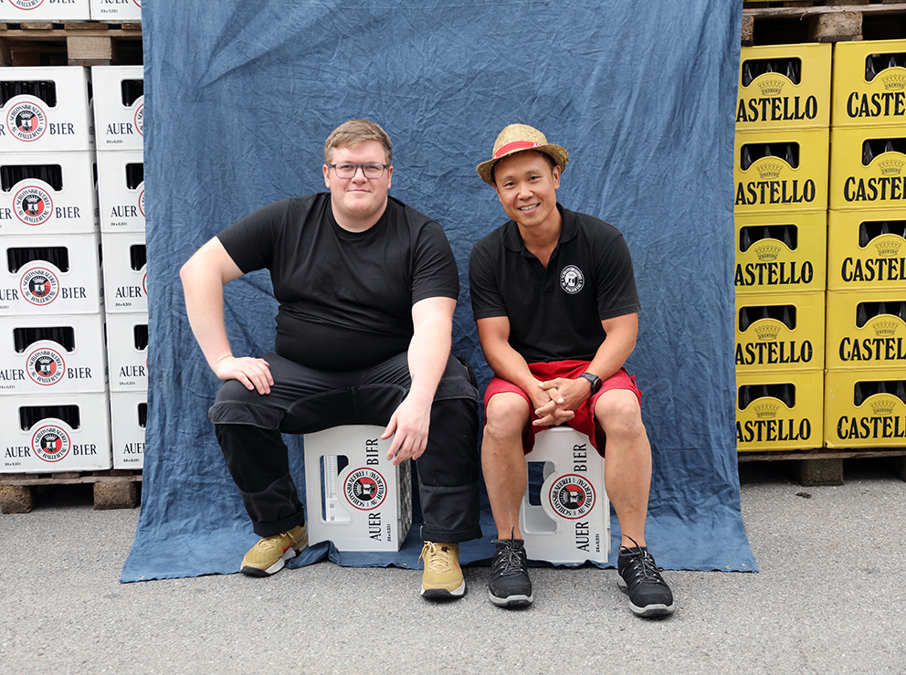 Two brewmasters seated side by side on Auer Bier crates in front of a blue backdrop