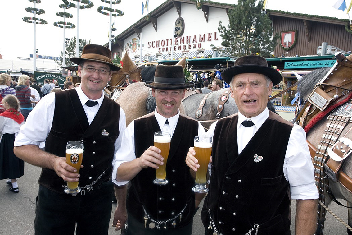 Three men in traditional Bavarian dress raising glasses of beer beside brewery horses outside the Schottenhamel tent