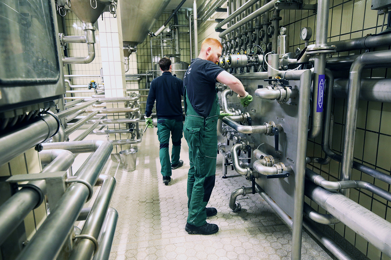 Two Barre brewery workers in green overalls working among stainless steel pipes and machinery