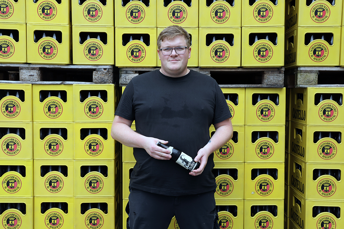 A brewer holding a bottle of Auer Helles in front of stacked yellow Schlossbrauerei crates