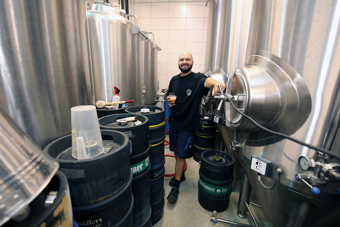 A brewer standing among kegs and tanks holding a glass of beer