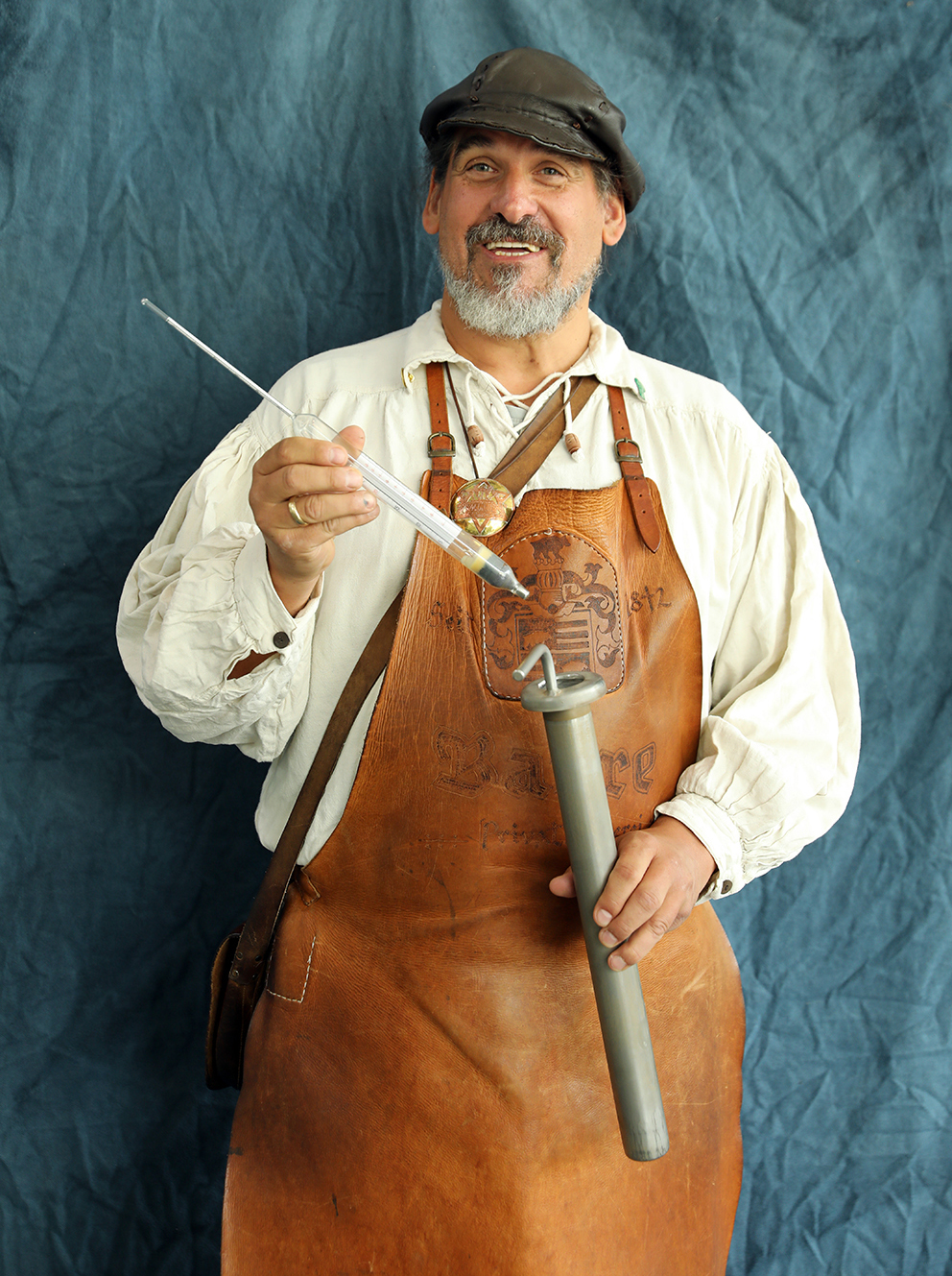 A bearded brewer in a traditional leather apron holding a hydrometer and sampling tube