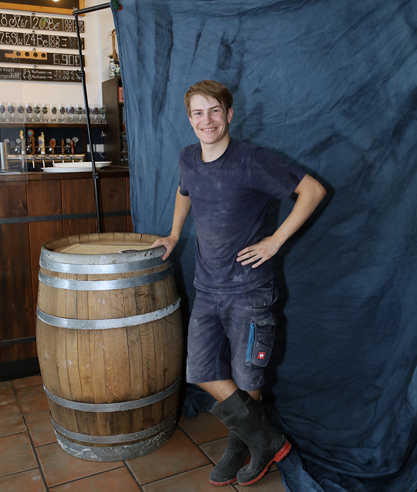 A young brewer in working clothes standing beside a wooden barrel