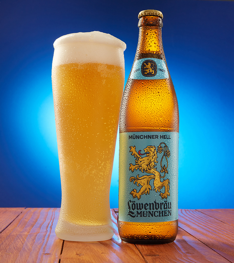 Löwenbräu München bottle and tall Weissbier glass on a wooden table with blue backdrop