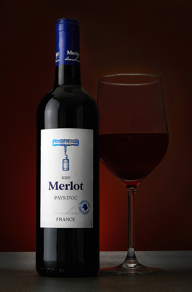 A Merlot Pays d'Oc bottle beside a glass of red wine on a warm red backdrop