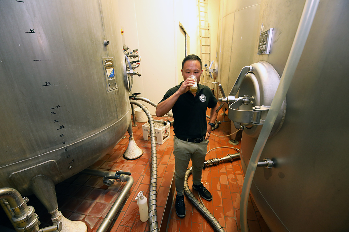 A brewer tasting beer from a glass between stainless steel fermentation tanks