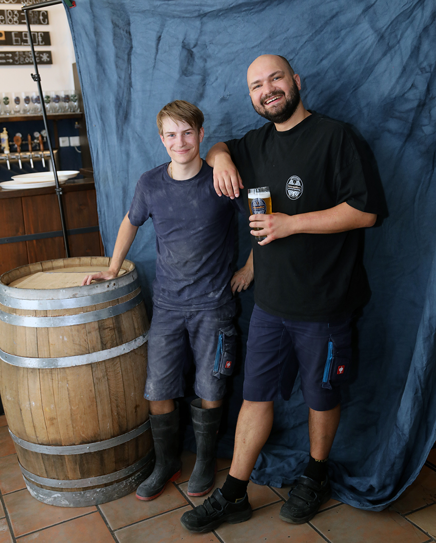 Two brewers standing beside a wooden barrel, one holding a glass of beer