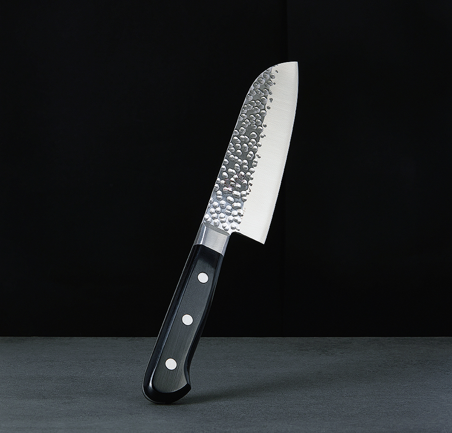 Angled Japanese chef's knife with hammered blade and dark handle on a grey surface