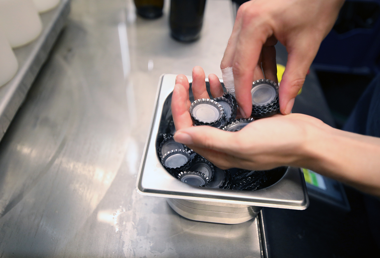 Hands lifting crown caps out of a stainless steel tray beside the bottling line